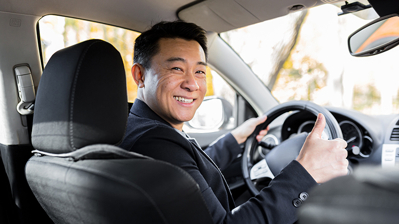 A man driving an Asian car behind the wheel looks at the camera and smiles